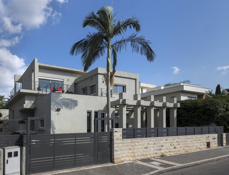 מבט חזיתי על הבית הפרטי לאחר השיפוץ וההרחבה ברמת ישי, בתכנון אדריכלי מודרני המאופיין בקווים נקיים, מסות גיאומטריות וחומריות מינימליסטית בגווני אפור בהיר. חזית הבית משלבת מרפסות פתוחות, פרגולה קונסטרוקטיבית ושפה אחידה של גדרות ופתחים כהים היוצרים קונטרסט אלגנטי למעטפת הבהירה. עץ הדקל הגבוה במרכז החזית מוסיף נוכחות טבעית ומרכך את הקווים האדריכליים הישרים, תוך יצירת תחושת יוקרה רגועה ואווירה ים־תיכונית עכשווית.